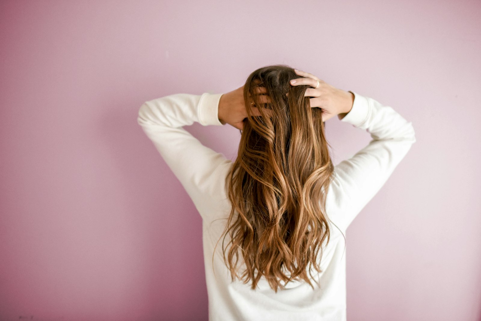A woman viewed from behind gathering long wavy hair against a pink wall