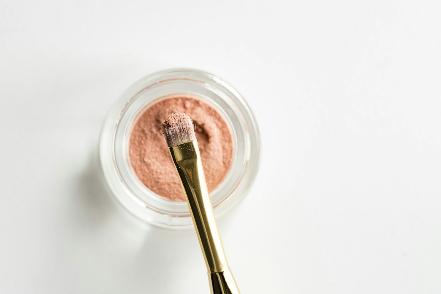 A gold fanned makeup brush resting on a pot of rose-toned cream on a white surface